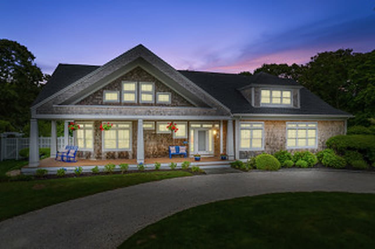 Gray-shingled home at twilight with every window lit, two blue chairs on the front porch, and a curved driveway across the lawn